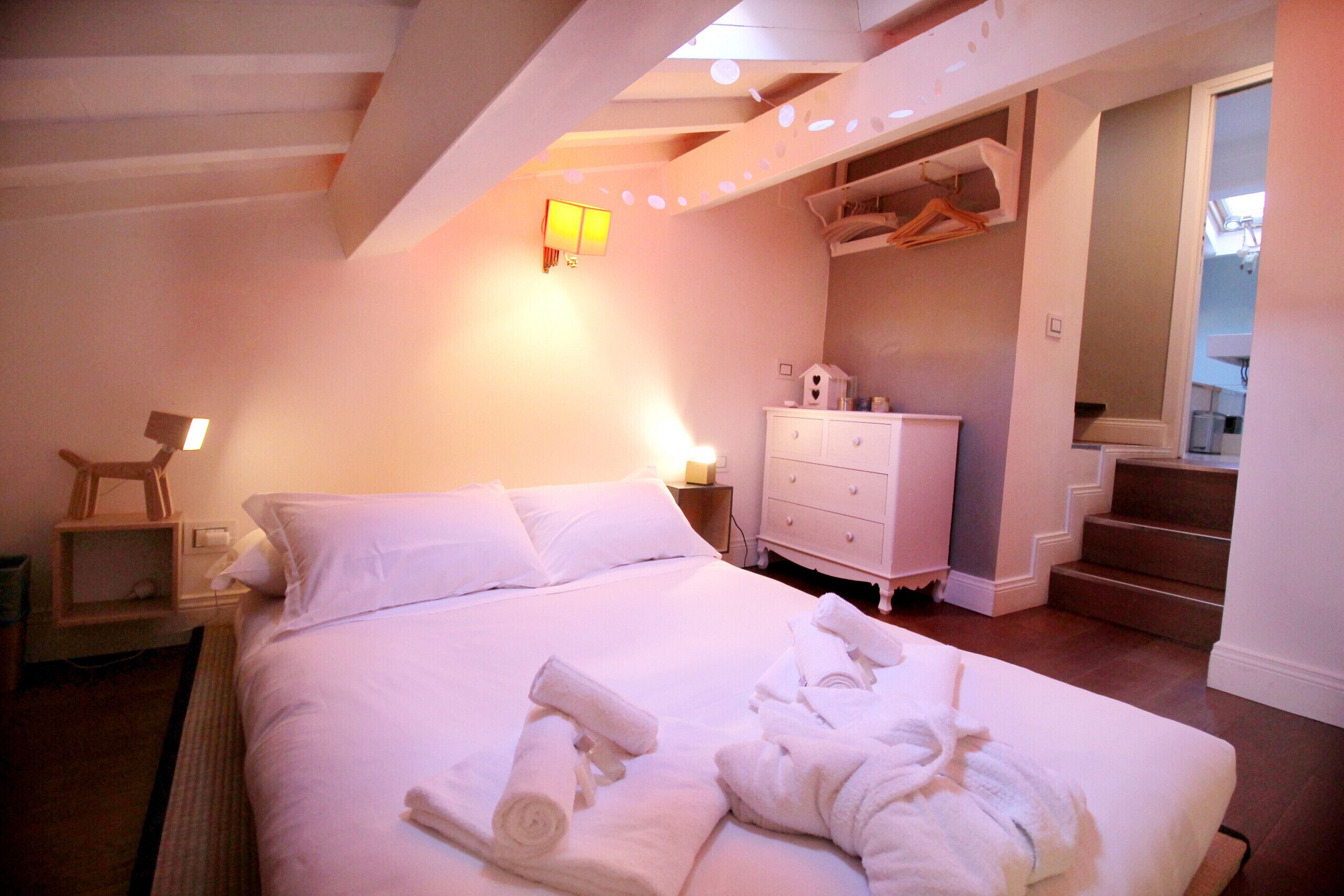 A neatly made bed with rolled towels, a bedside table with a lamp, a dresser, and a staircase leading to an upper level.