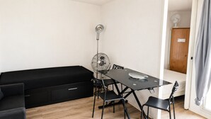 A compact room with a black sofa, a small table with chairs, a standing fan, and a door leading to another room. - Appartement 2 Pièces à Canet avec Vue Mer (Canet-en-Roussillon)