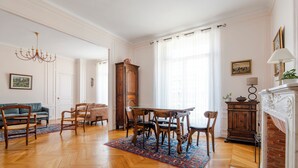 A spacious room with a wooden herringbone floor, a dining table with chairs, a chandelier, a fireplace, and a large window with sheer curtains.