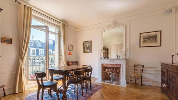 A dining room with a wooden table, chairs, and a patterned rug. There is a fireplace with a mantel and a mirror above it.
