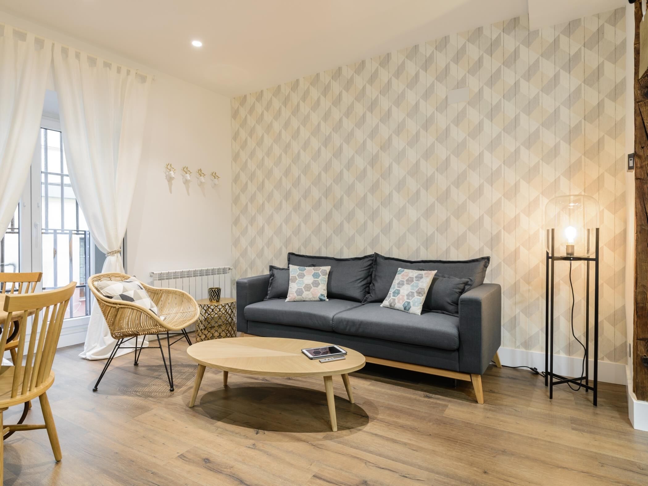 Un salón con un sofá gris, una mesa de centro redonda de madera, sillas de mimbre y un papel tapiz estampado.