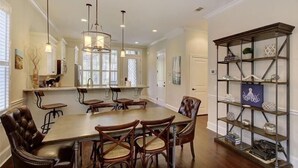 A dining area with a large wooden table and leather chairs, a chandelier, and a metal shelving unit with decorative items. - Garden Cottage 01 - The Cottages at Jekyll Island oceanfron community. (Jekyll Island)