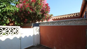 A covered patio area with a white gate, a red wall, and a tree with pink flowers. - Maisonette T2  et parking dans résidence avec piscine (Canet-en-Roussillon)