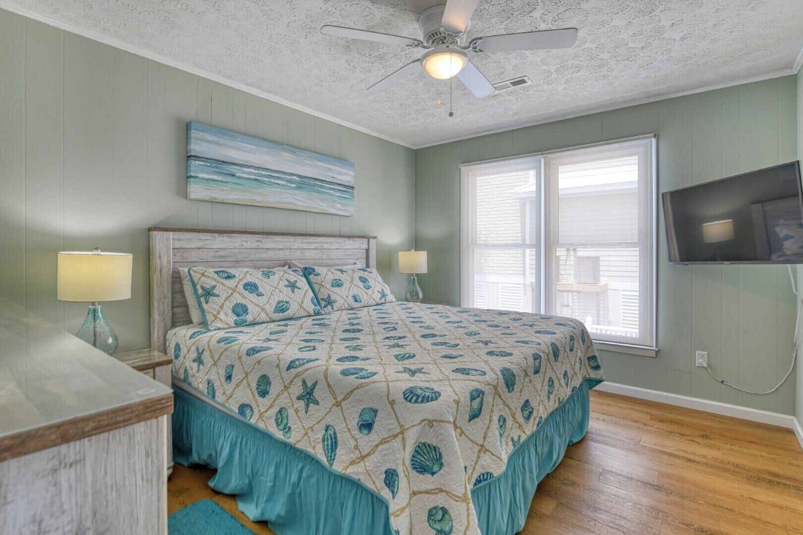 A bedroom with a bed, two bedside lamps, a ceiling fan, a TV mounted on the wall, and a window with blinds.