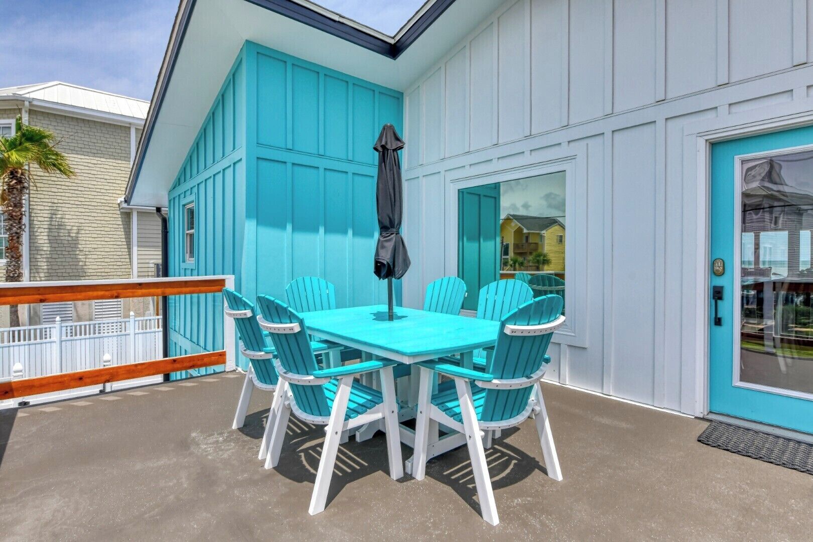Un espace extérieur aménagé pour les repas, avec une table et des chaises, un parasol noir et une rambarde blanche, le tout dans des tons turquoise.