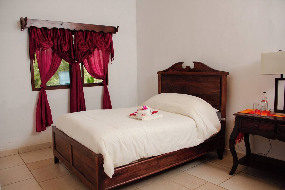 A neatly made bed with a white comforter and pillows, a wooden headboard, and a window with red curtains.