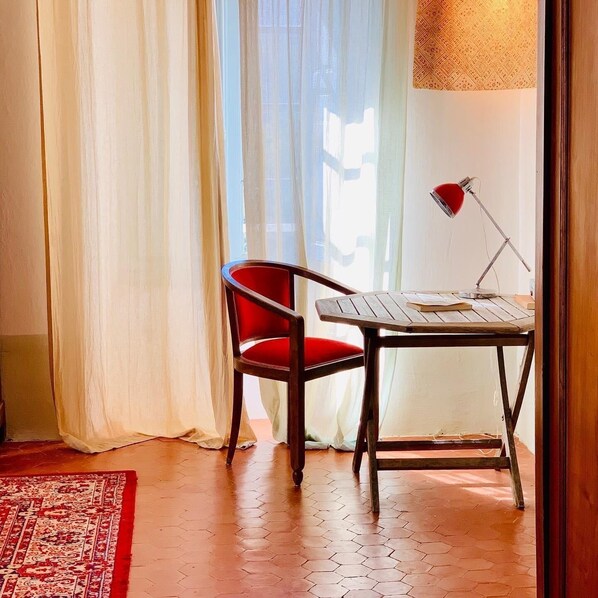 Una habitación con una mesa de madera, una silla roja, un tapete estampado y una ventana con cortinas de encaje.