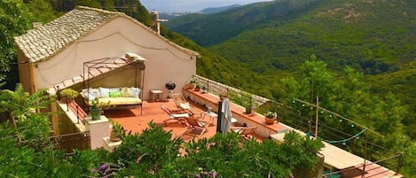 A rustic villa with a terracotta roof, a covered outdoor seating area, and a scenic mountainous backdrop.