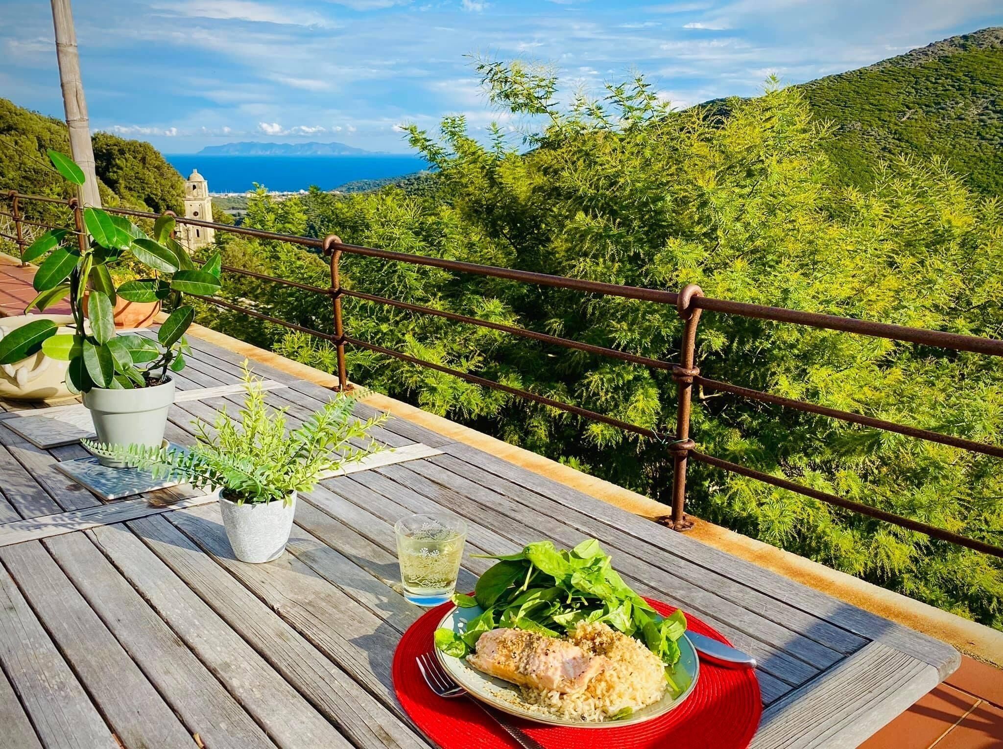 Une terrasse en bois avec une table dressée pour un repas, offrant une vue imprenable sur un paysage verdoyant et une perspective lointaine sur la mer.