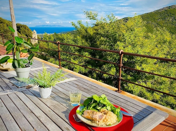 A wooden deck with a table set for a meal, overlooking a lush green landscape and a distant view of the sea.