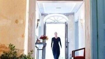 Un couloir dans lequel une personne marche en direction d’une porte. Il y a une plante en pot sur la gauche et une chaise sur la droite.