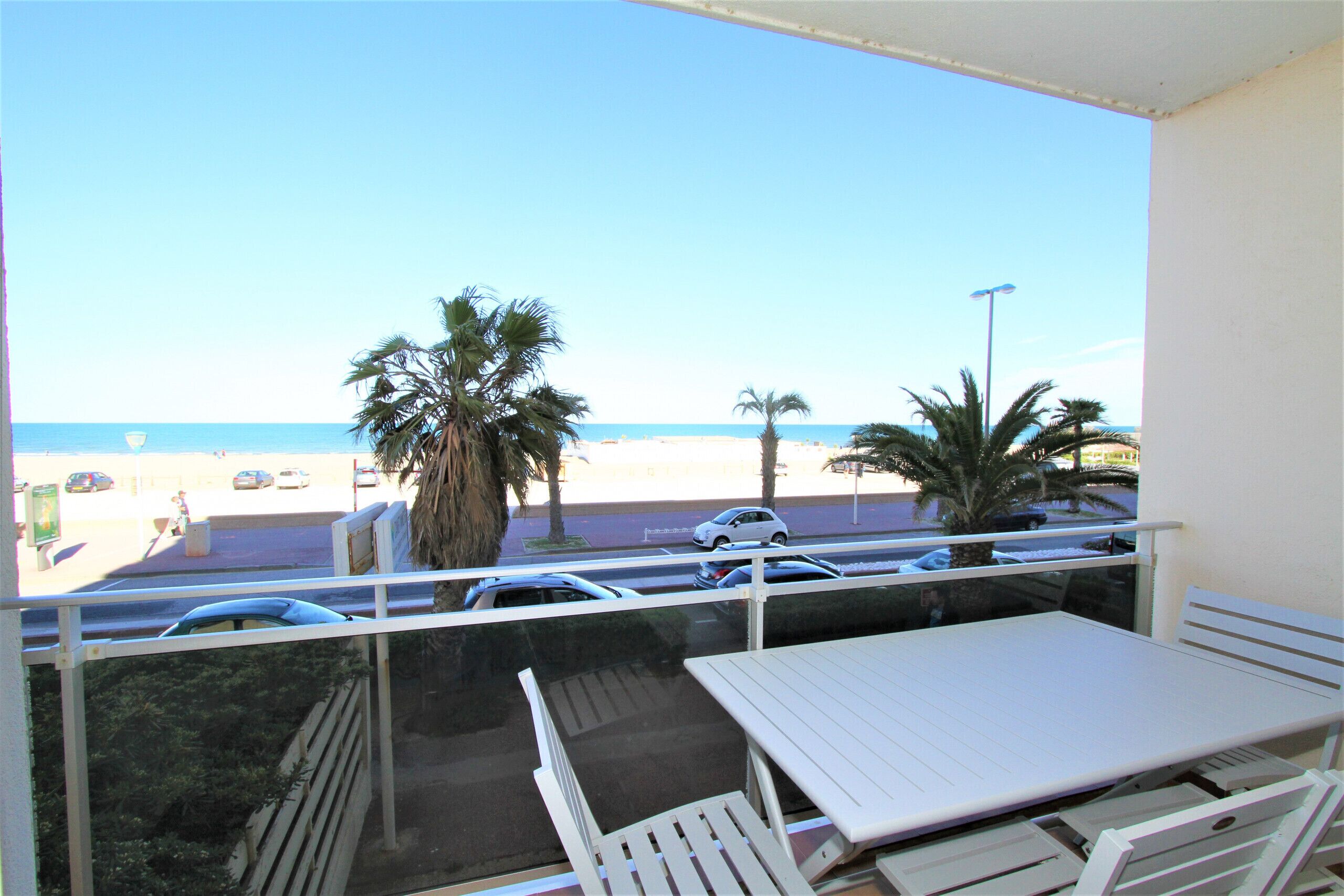 A balcony with a table and chairs, overlooking a beach with palm trees and parked cars.