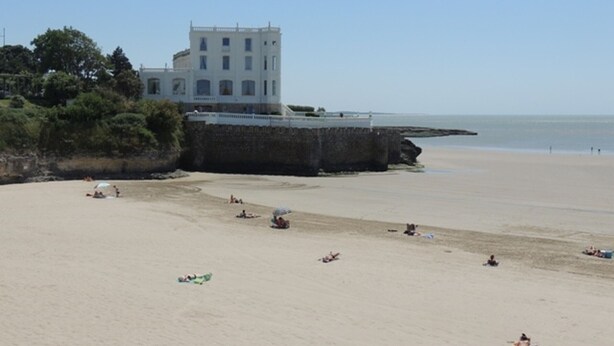 A beach with a large building in the background, a stone wall, and a clear sky.