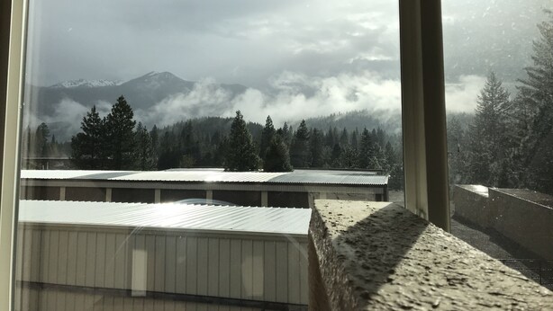 Vista a través de una ventana de un paisaje montañoso con picos nevados, una zona boscosa y un edificio con techo de metal corrugado.