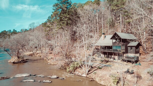 A cabin with a balcony overlooking a river, surrounded by a forest.