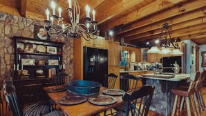 A rustic wooden dining area with a stone fireplace, a chandelier, and a kitchen in the background.