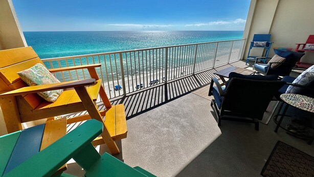 Een balkon met uitzicht op zee, houten stoelen, een tafel en een zitje met kussens.