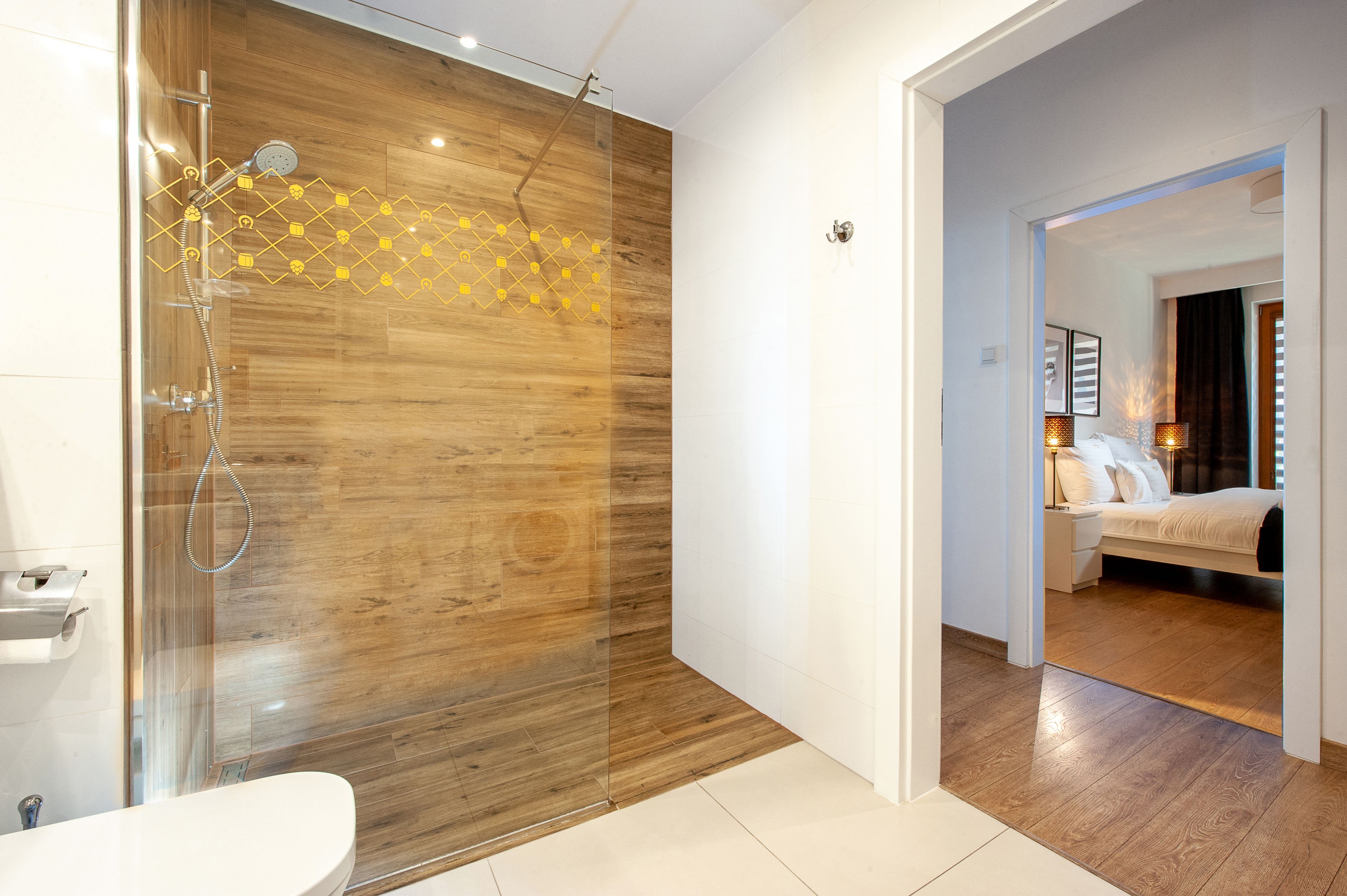 Un baño moderno con ducha envuelta en vidrio, paneles de madera, inodoro blanco y una habitación visible con cama y mesita de noche.