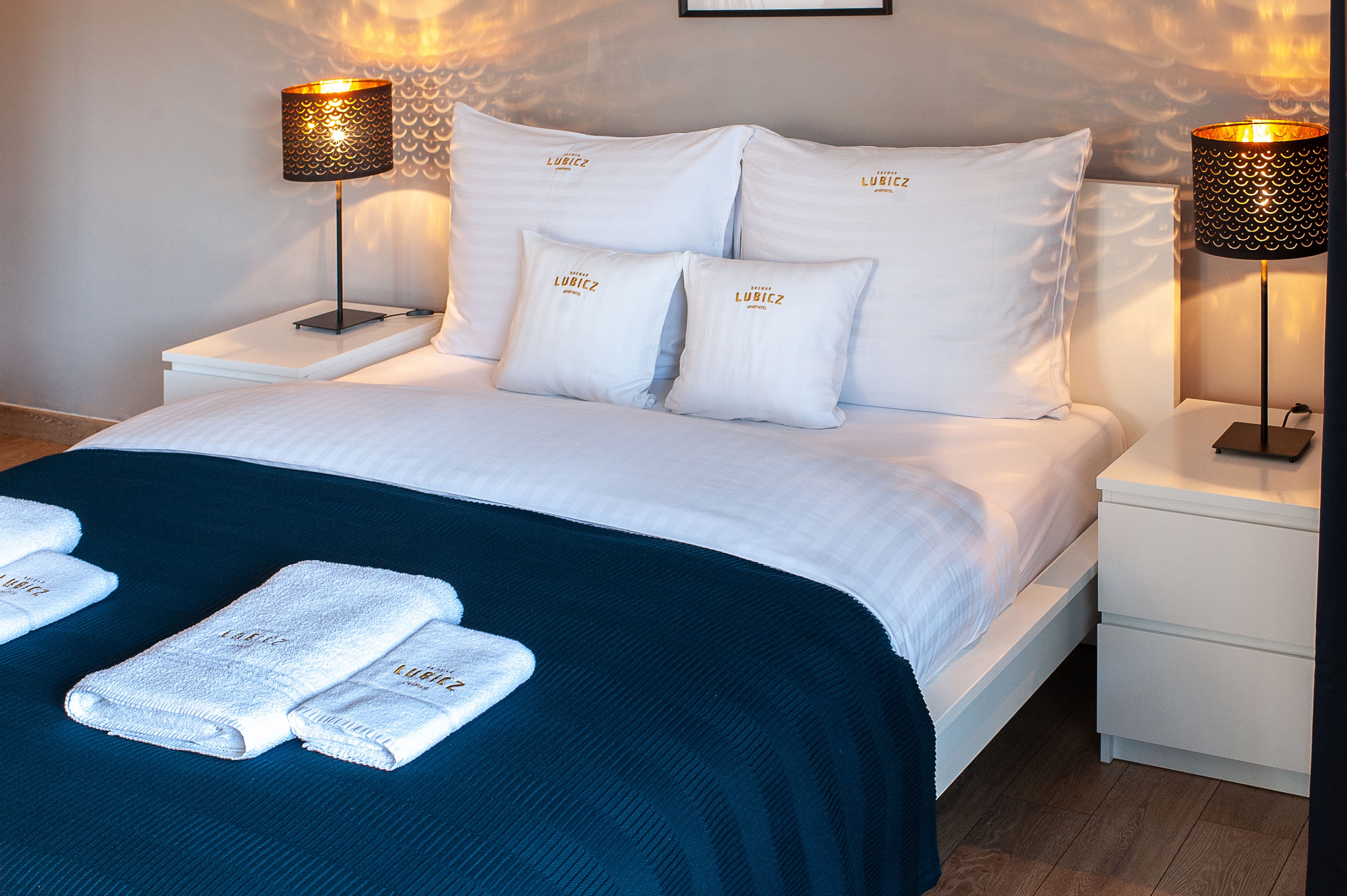A neatly made bed with white linens and pillows, two bedside lamps, and two folded towels on the bed.