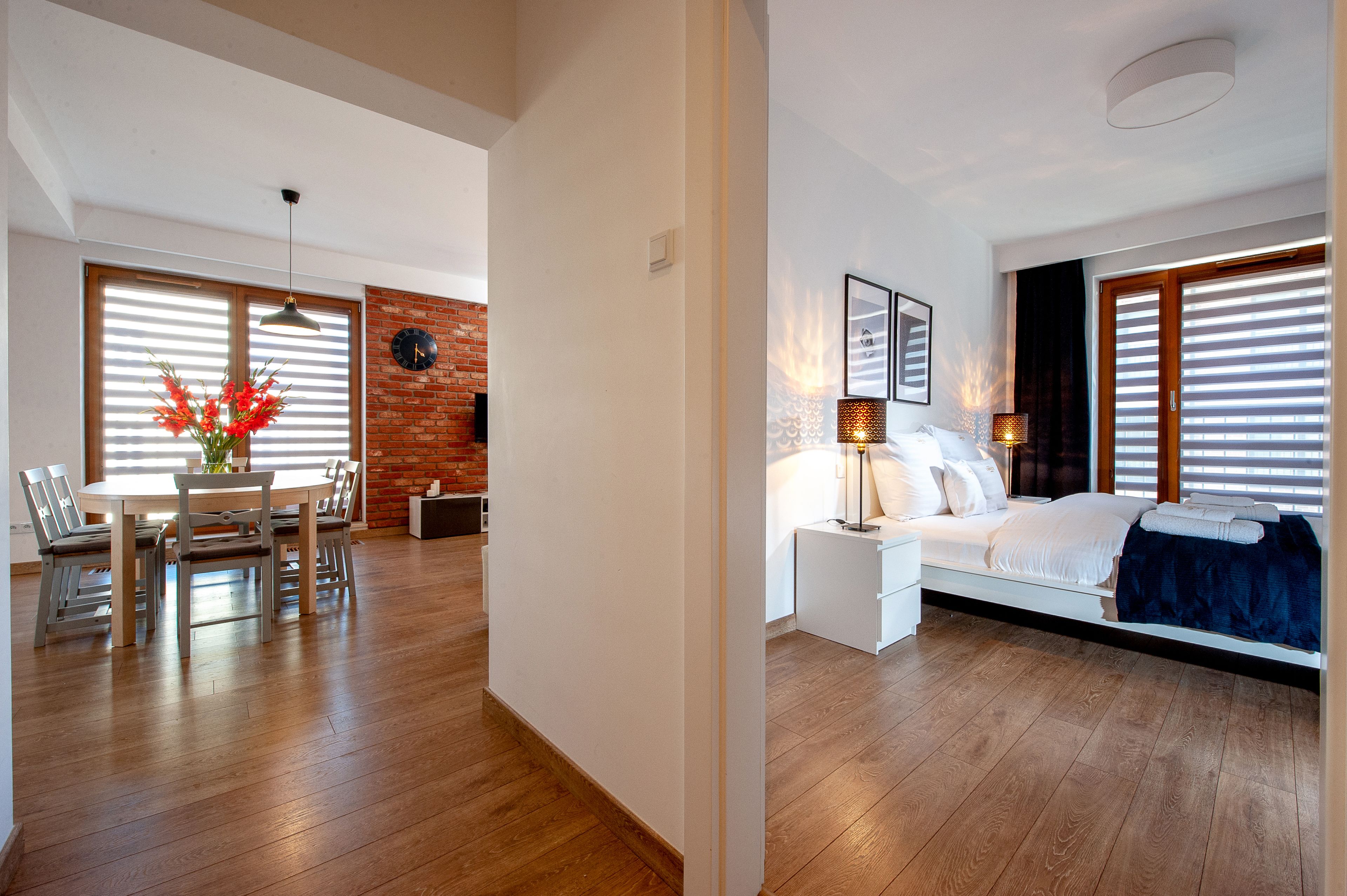 A modern hotel room with a wooden floor, a bed with white bedding, a bedside table with a lamp, and a view through large windows with blinds.