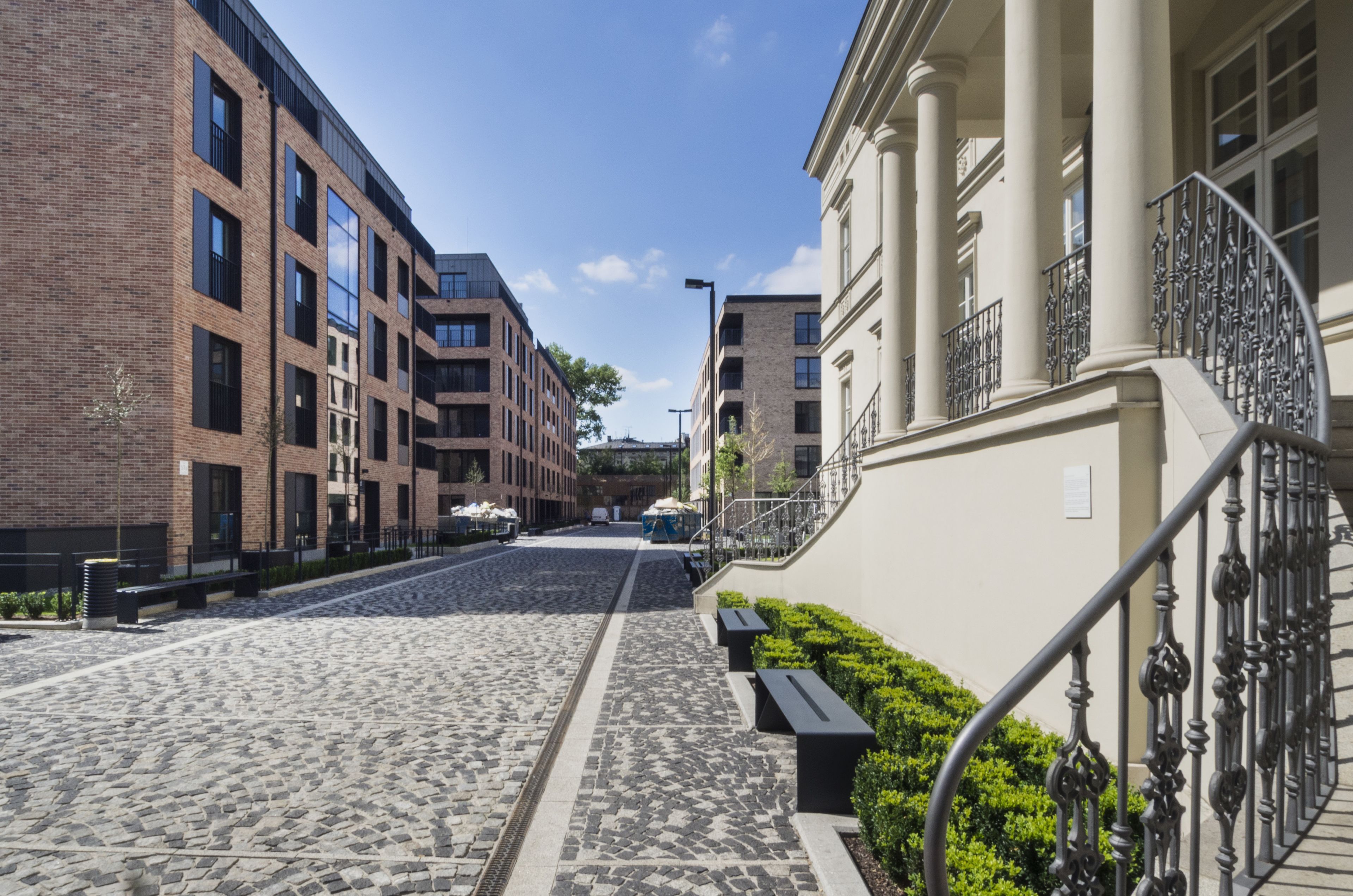 A cobblestone street lined with modern buildings, a staircase with ornate metal railings, and benches along the sidewalk.