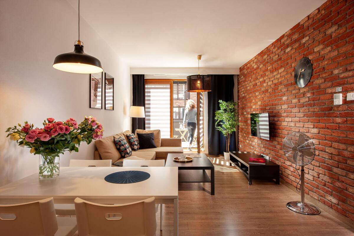 Una sala moderna con pared de ladrillo, un comedor con mesa y sillas, un sofá con cojines, un ventilador de pie y un ventanal grande.