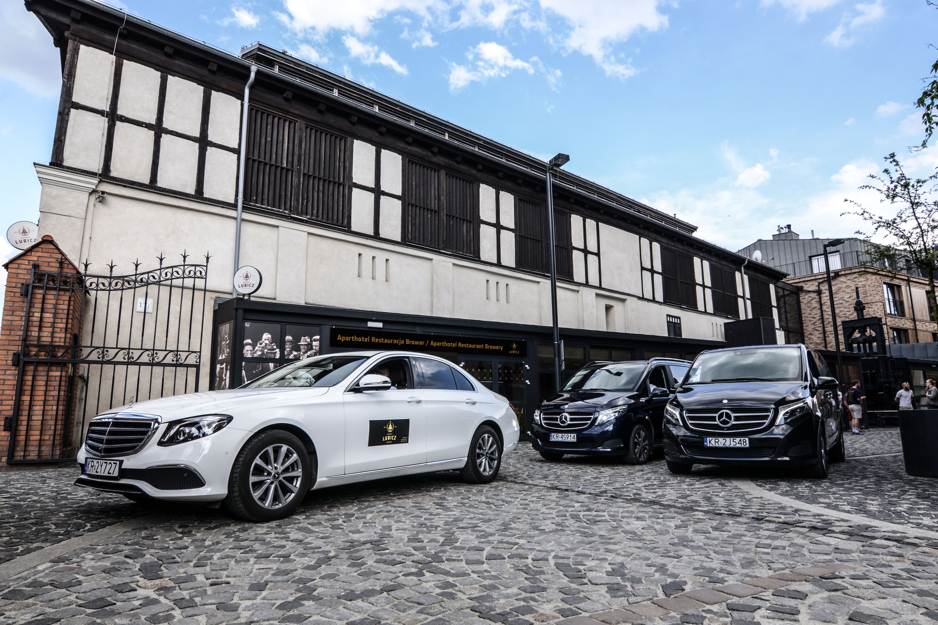 A cobblestone street with two parked cars, a building with a sign for an apartment hotel and restaurant, and a brick wall with a gate.