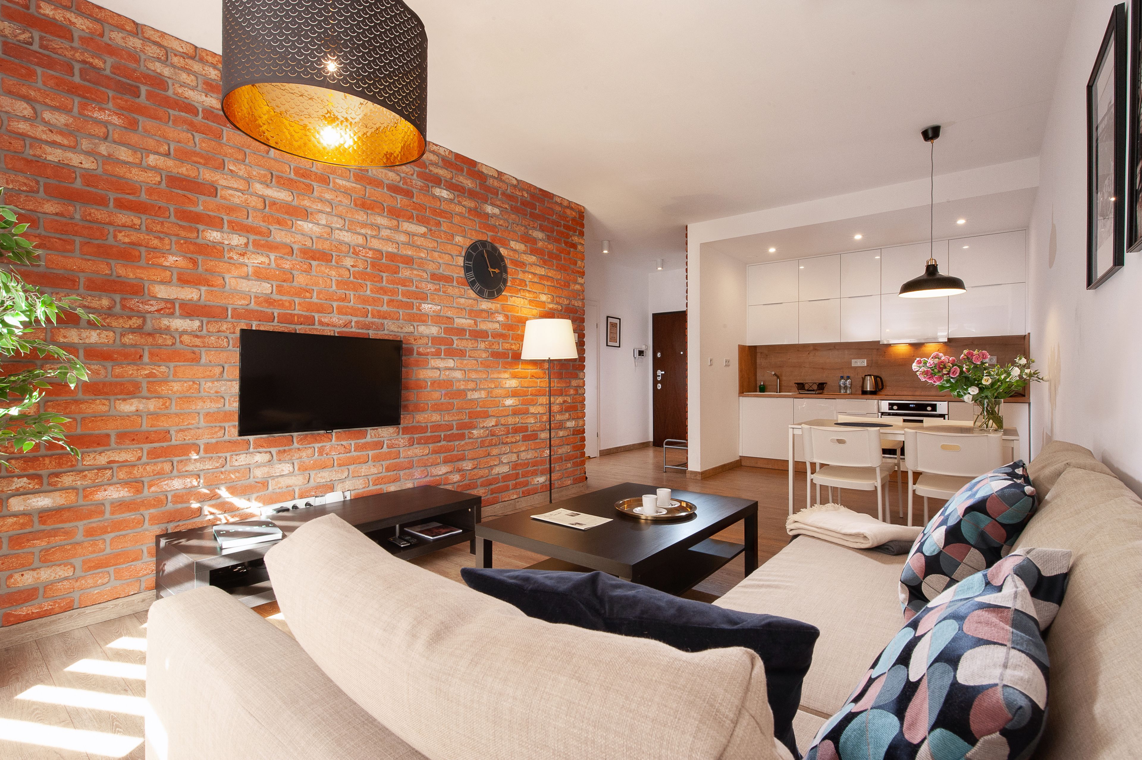 A modern living room with a brick wall, a flat-screen TV, a sofa, a coffee table, and a dining area with a kitchen in the background.