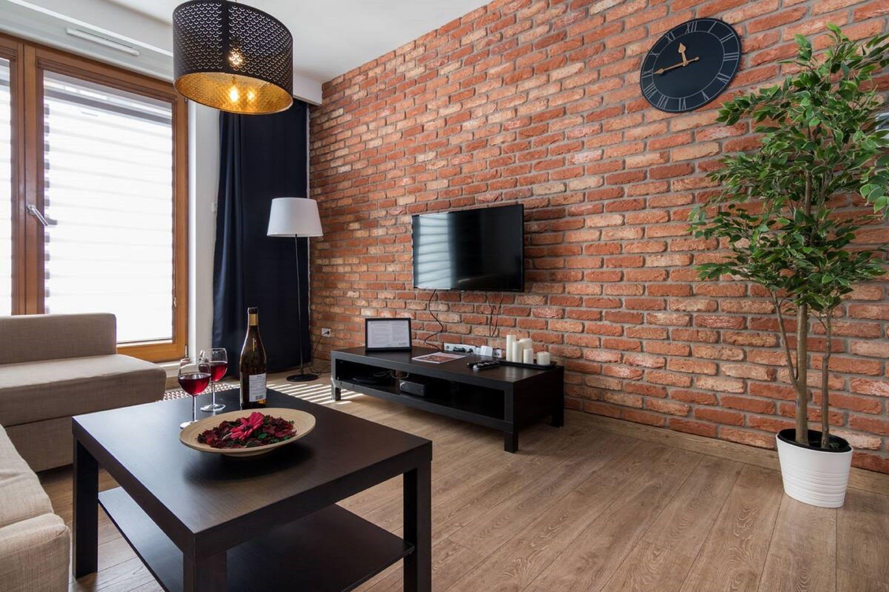 A modern living room with a brick wall, a flat-screen TV, a coffee table with a bowl of fruit, and a potted plant.