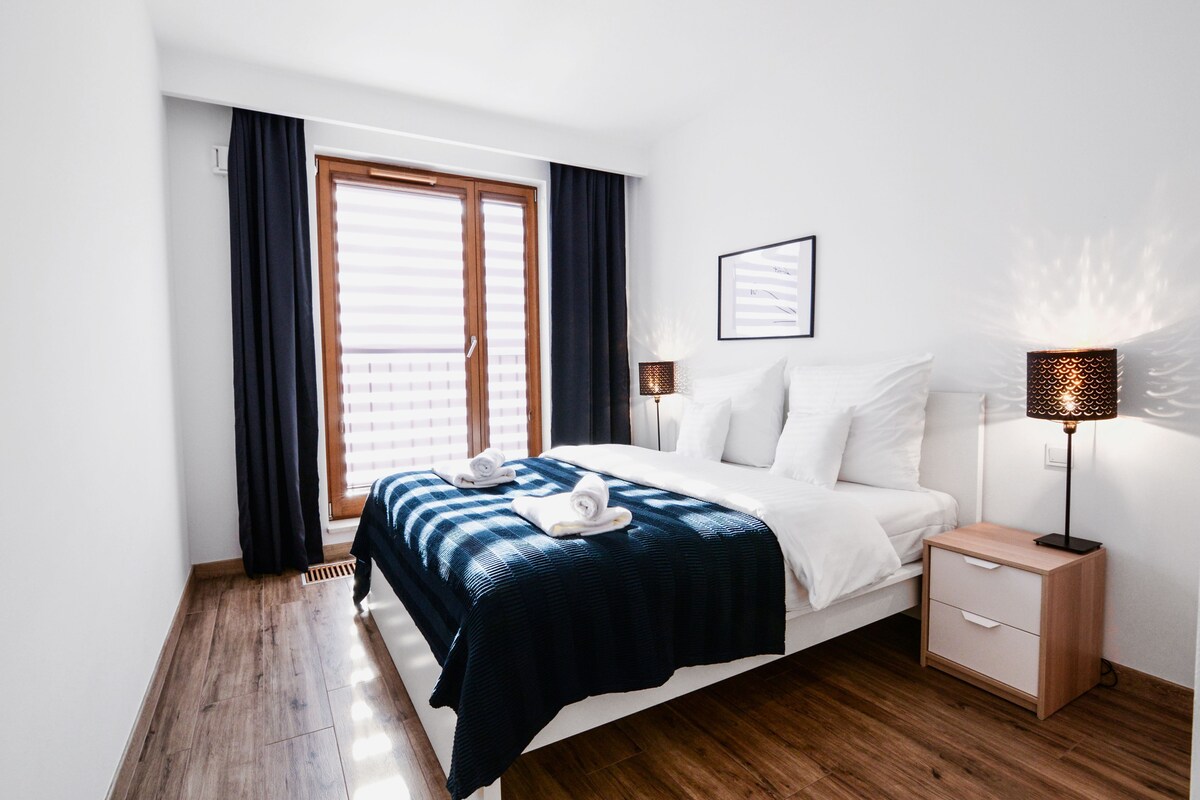 A modern bedroom with a large bed, two bedside tables, a nightstand, a window with curtains, and a wall-mounted lamp.