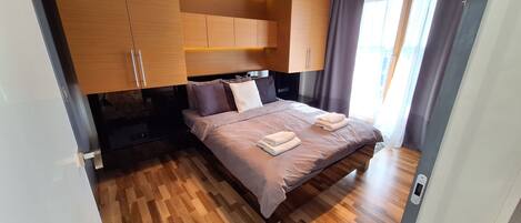 A modern bedroom with a wooden floor, a bed with a headboard, and a ceiling light fixture.