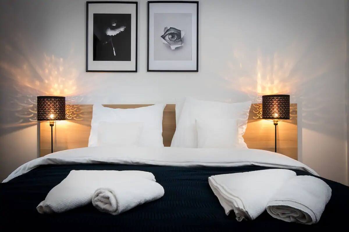 Una cama bien tendida con sábanas blancas, dos cuadros enmarcados en la pared y dos lámparas a cada lado.