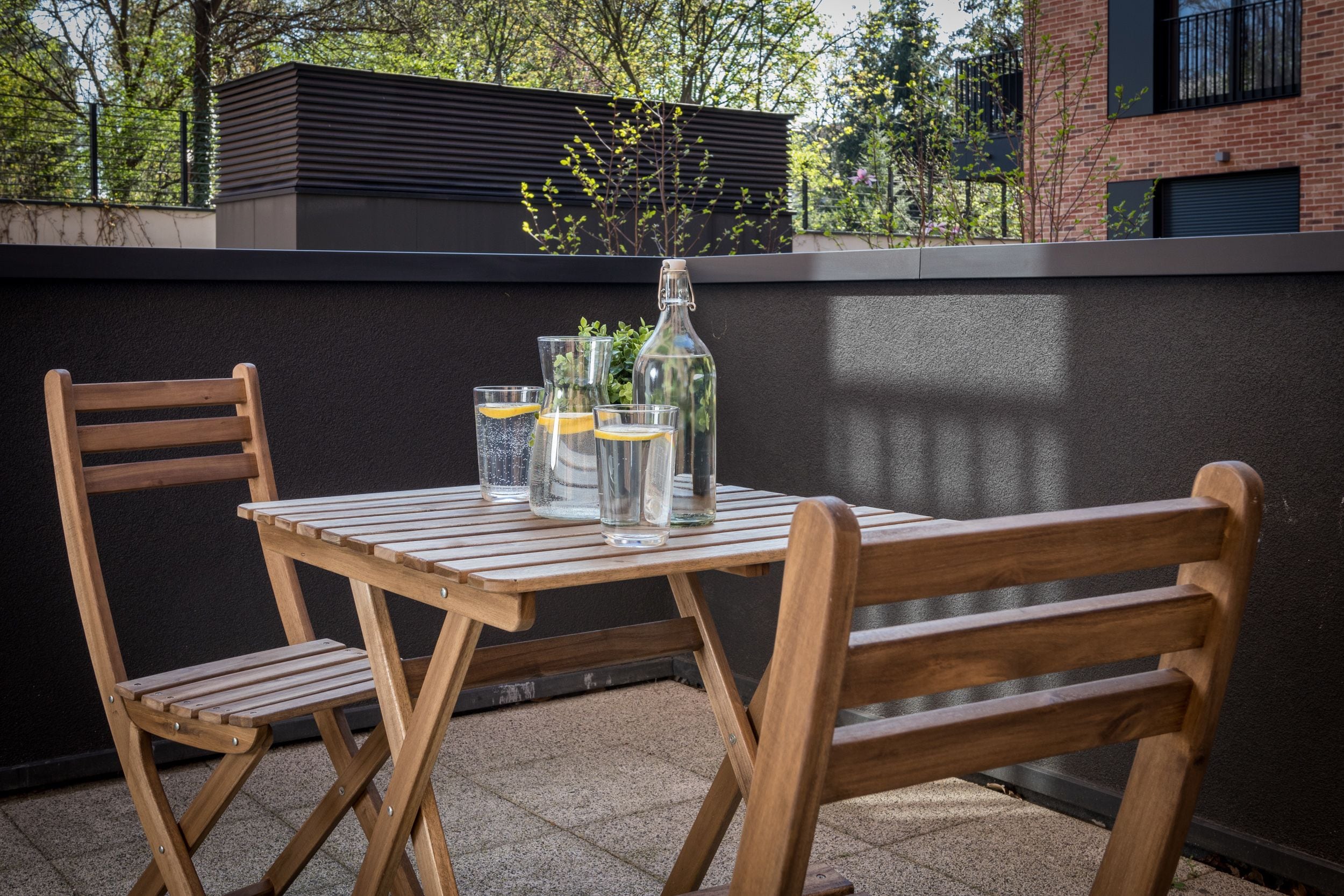 A wooden table and chairs on a balcony with a view of trees and a brick building.