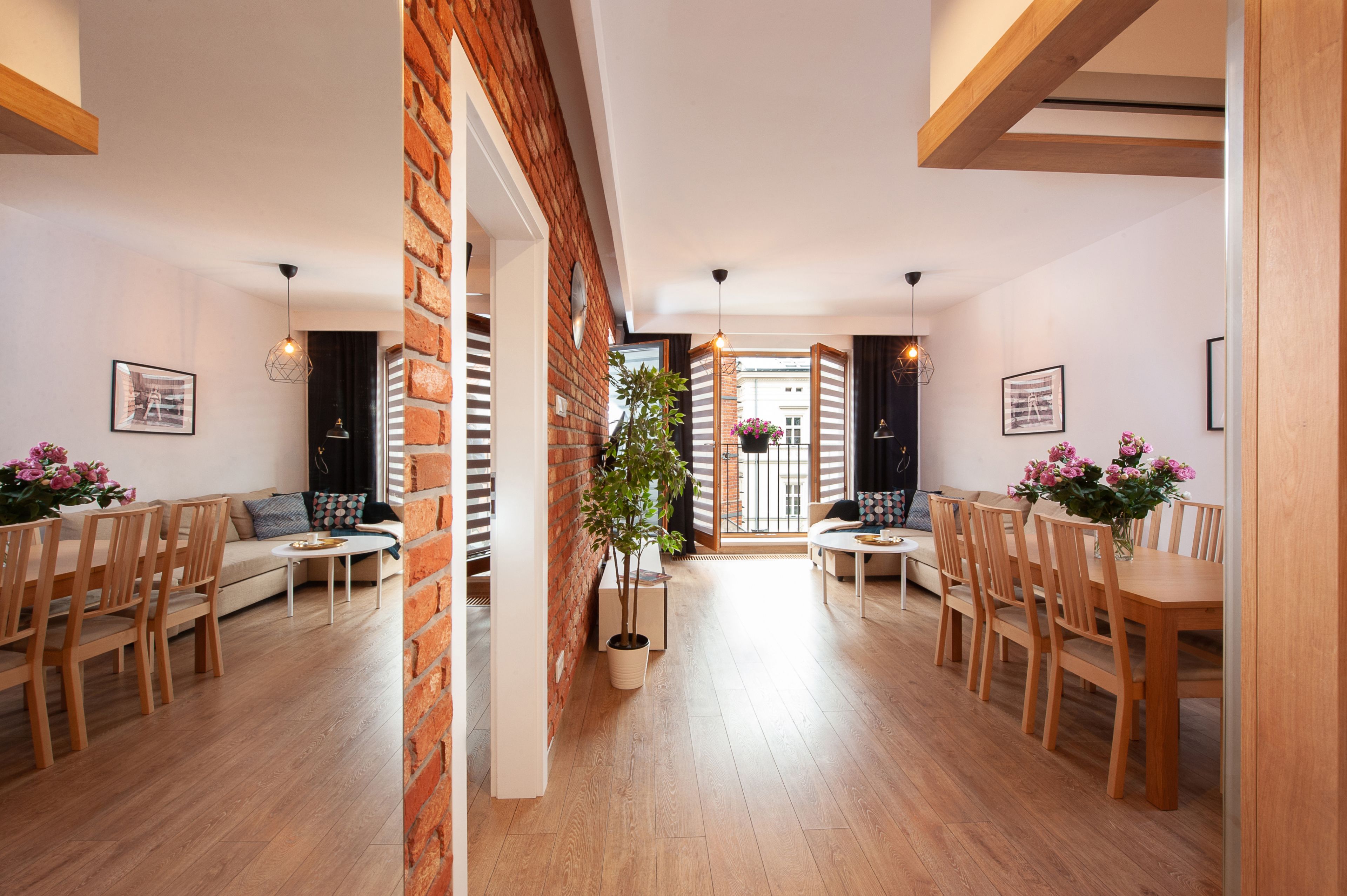 A spacious living area with a brick wall, wooden flooring, and a central plant. There are two seating areas with tables and chairs, and a staircase leading to another level.