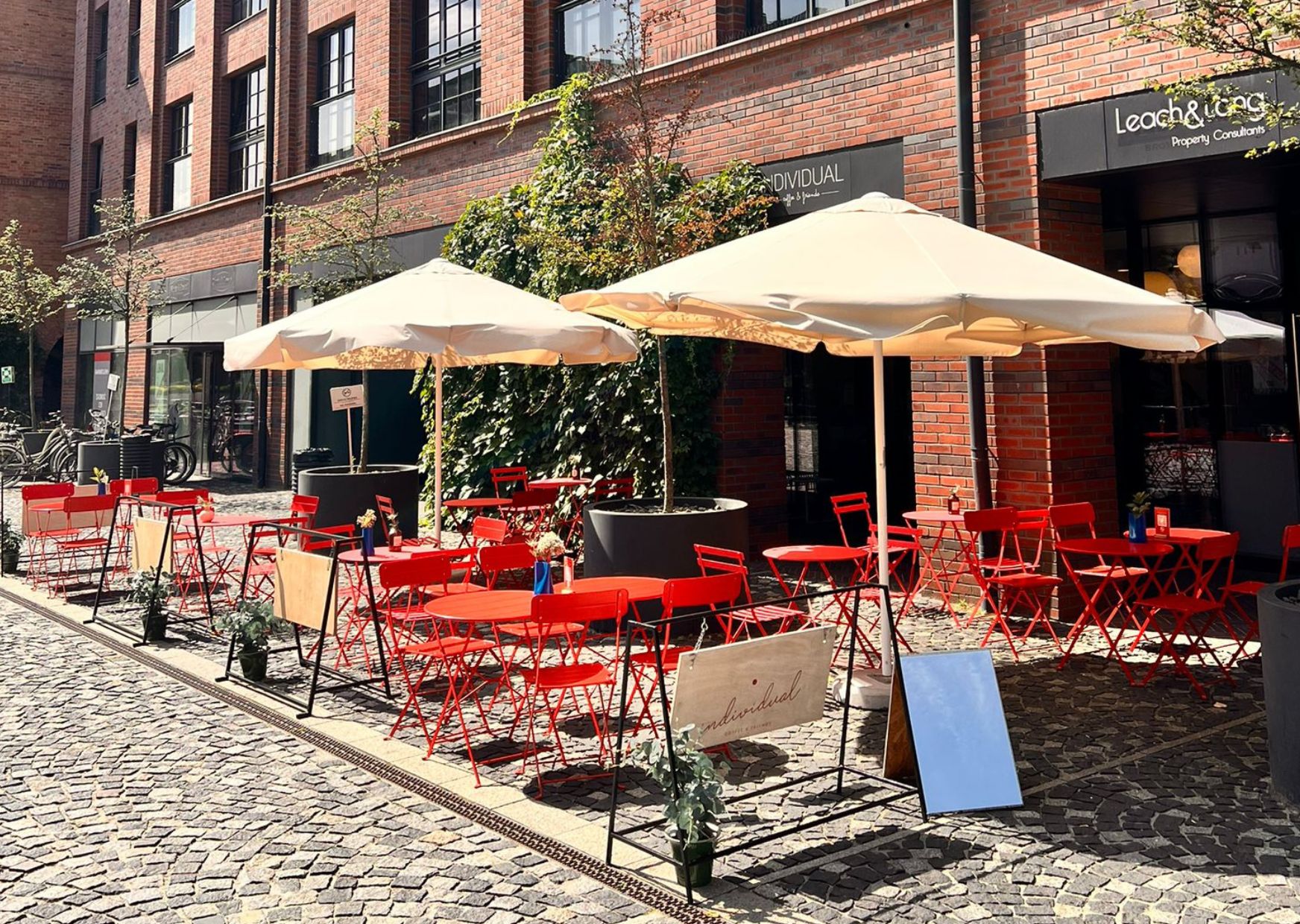An outdoor seating area with red chairs and tables, covered by beige umbrellas, in front of a brick building with a sign reading "Leach & Co."