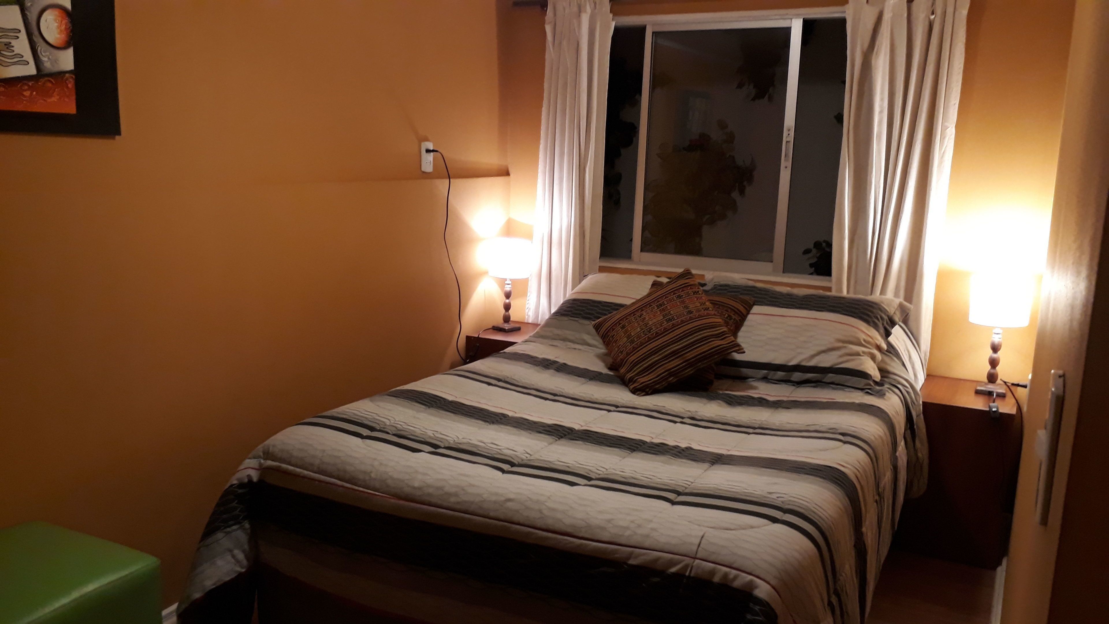 Una cama bien hecha con ropa de cama a rayas, dos lámparas de noche y una ventana con cortinas.