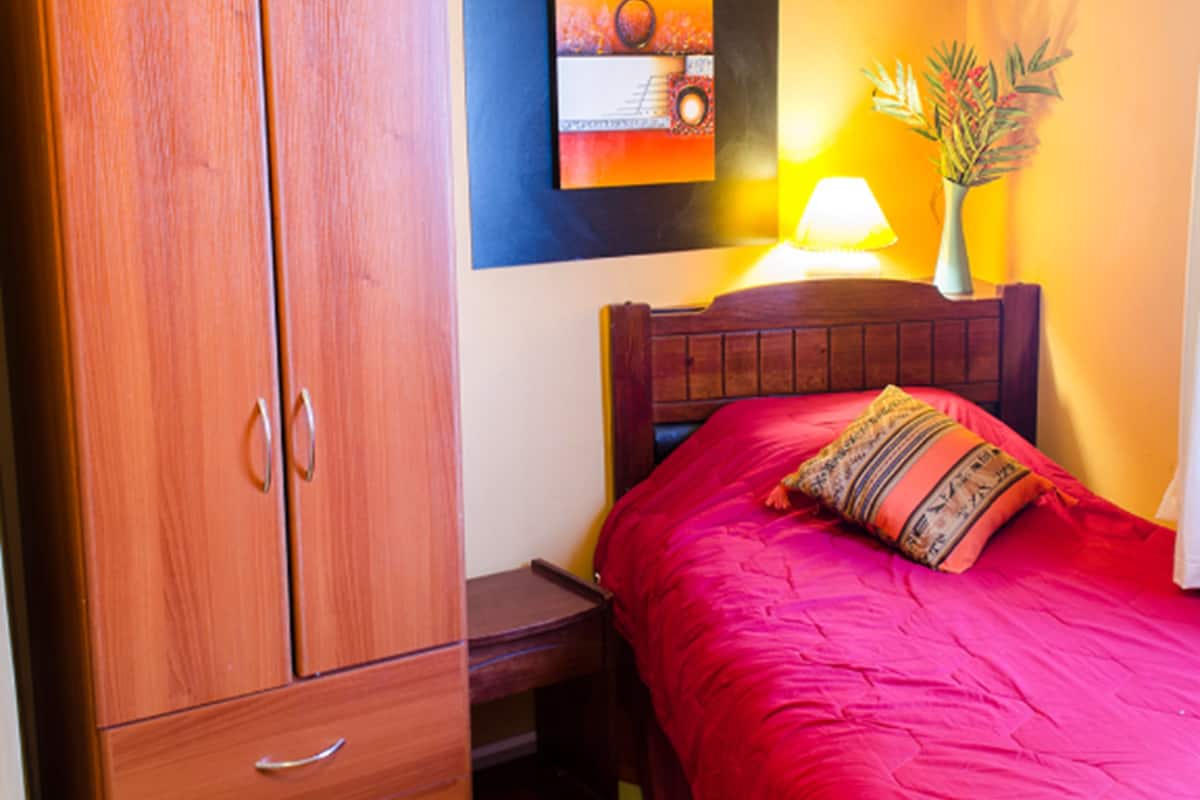 A bedroom with a wooden wardrobe, a bed with a red bedspread, a nightstand, a lamp, and a wall art piece.