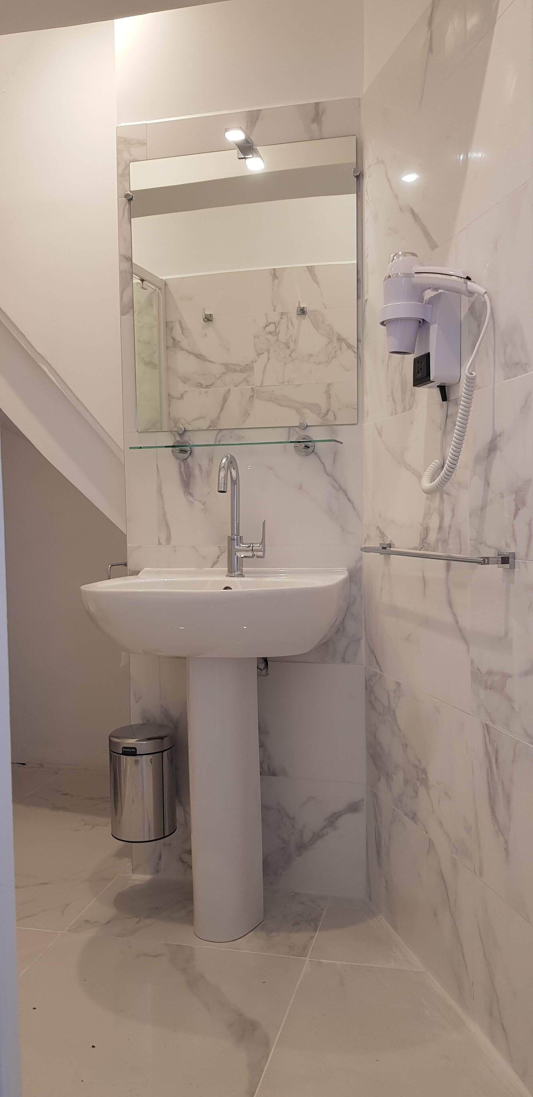 A modern bathroom with a marble sink, a glass shelf, a hairdryer, and a trash can.
