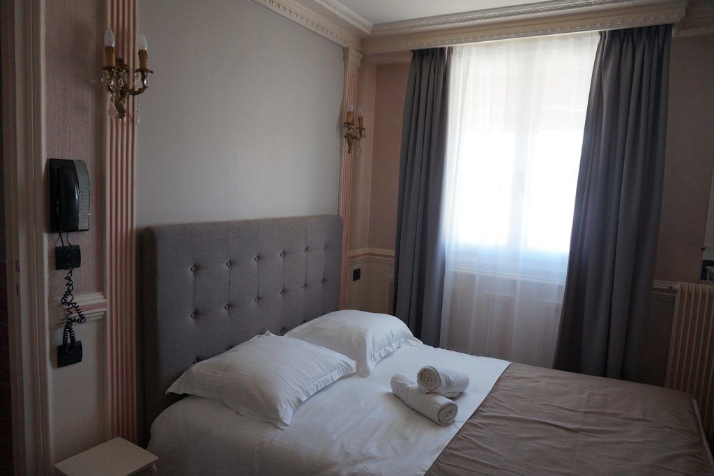 Una cama bien hecha con sábanas blancas y dos toallas enrolladas, un cabecero capitonado, un teléfono en la mesita de noche y una ventana con cortinas.