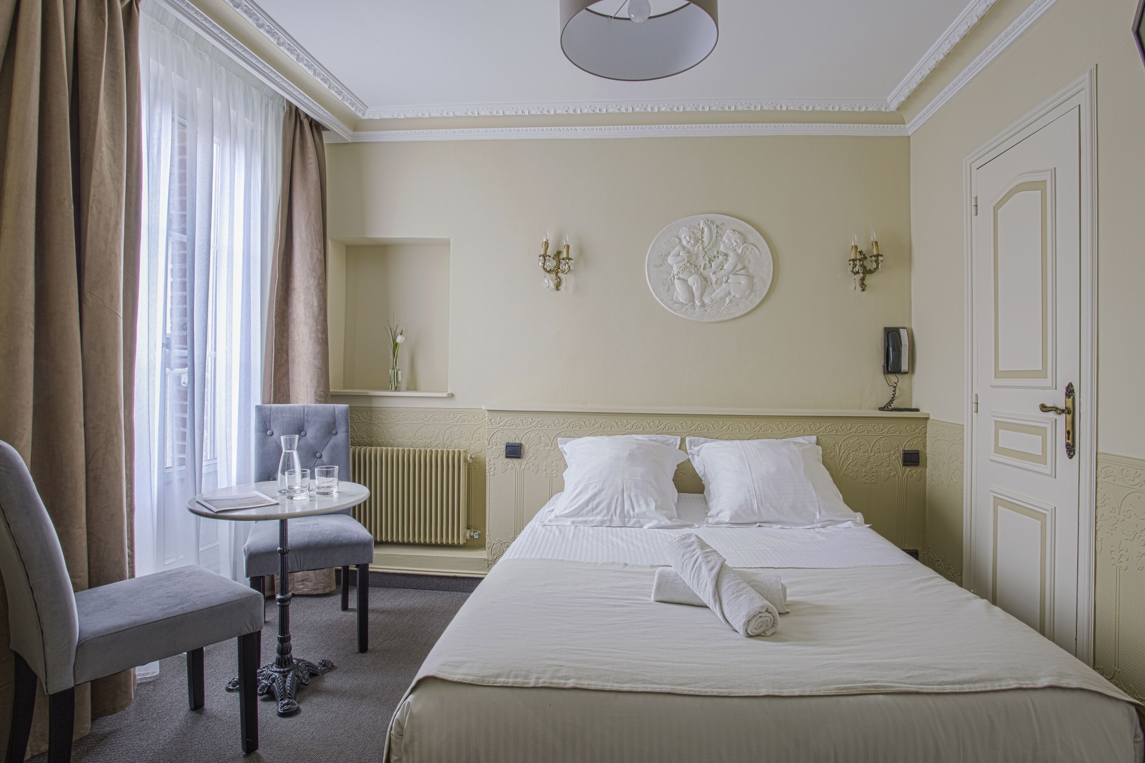 Habitación de hotel con una cama bien tendida, una mesita redonda con sillas, un radiador y una puerta con manija.