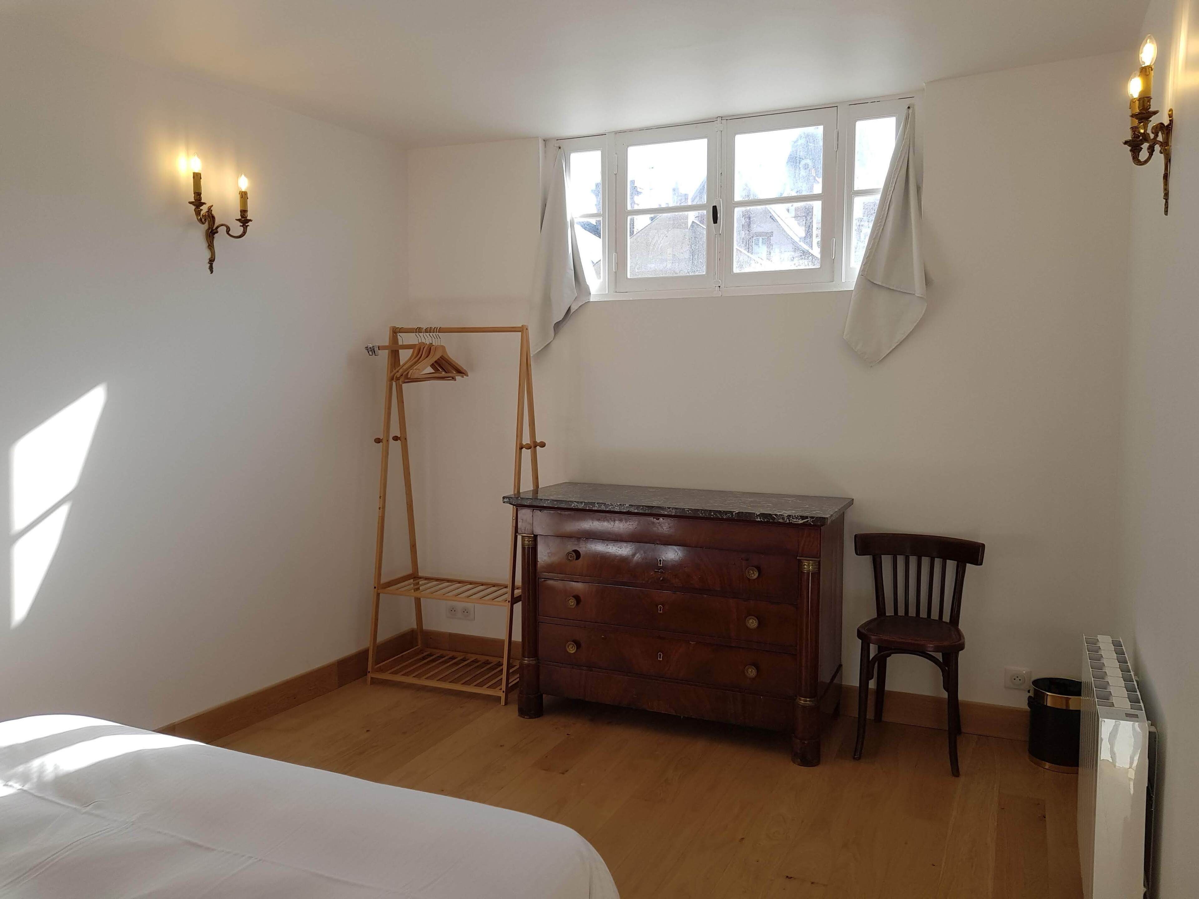 Un dormitorio con un aparador de madera, una silla, un tendedero de madera y una ventana con cortinas.