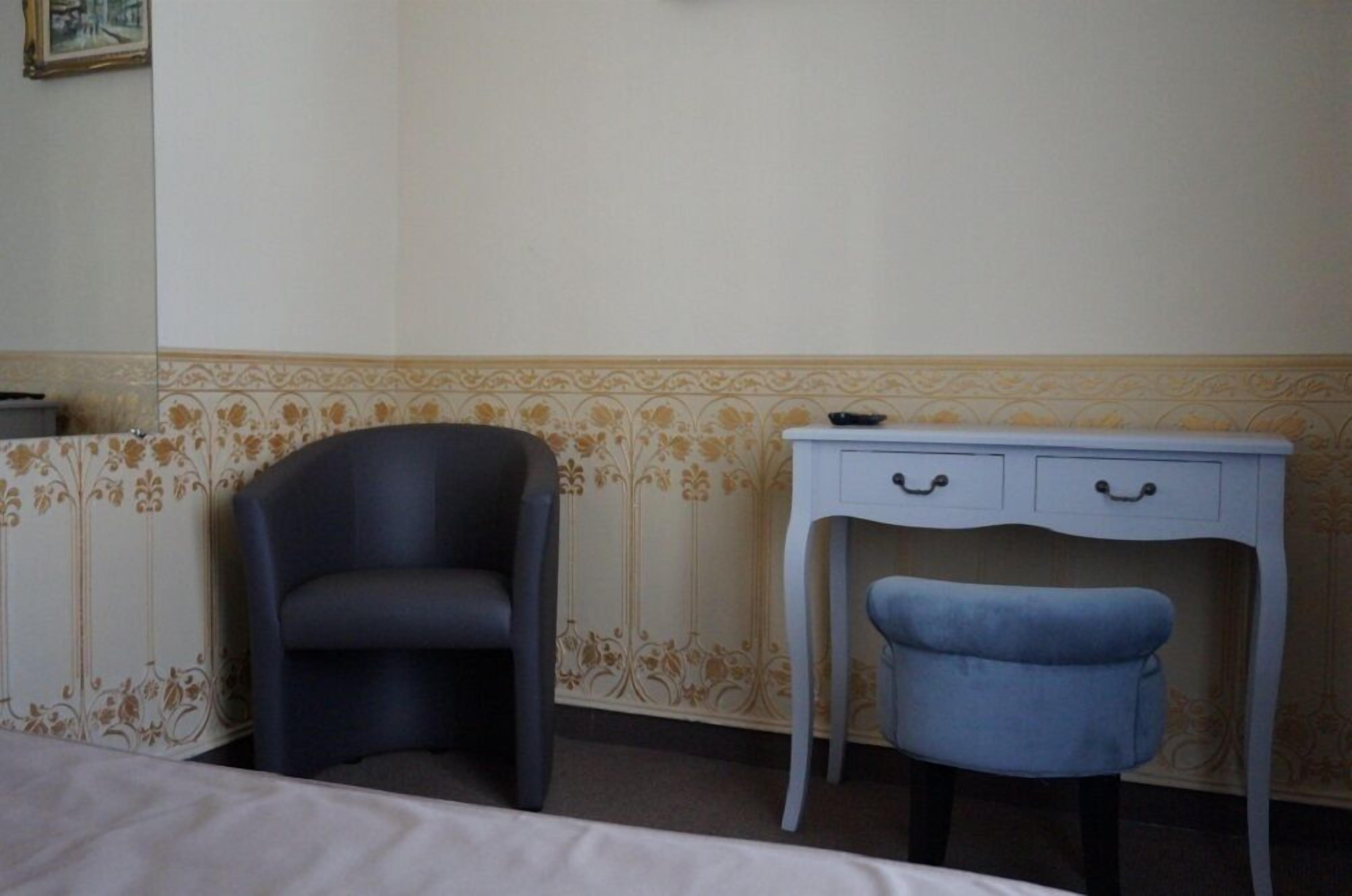 A room with a bed, a dark chair, a small desk with drawers, and a blue stool.