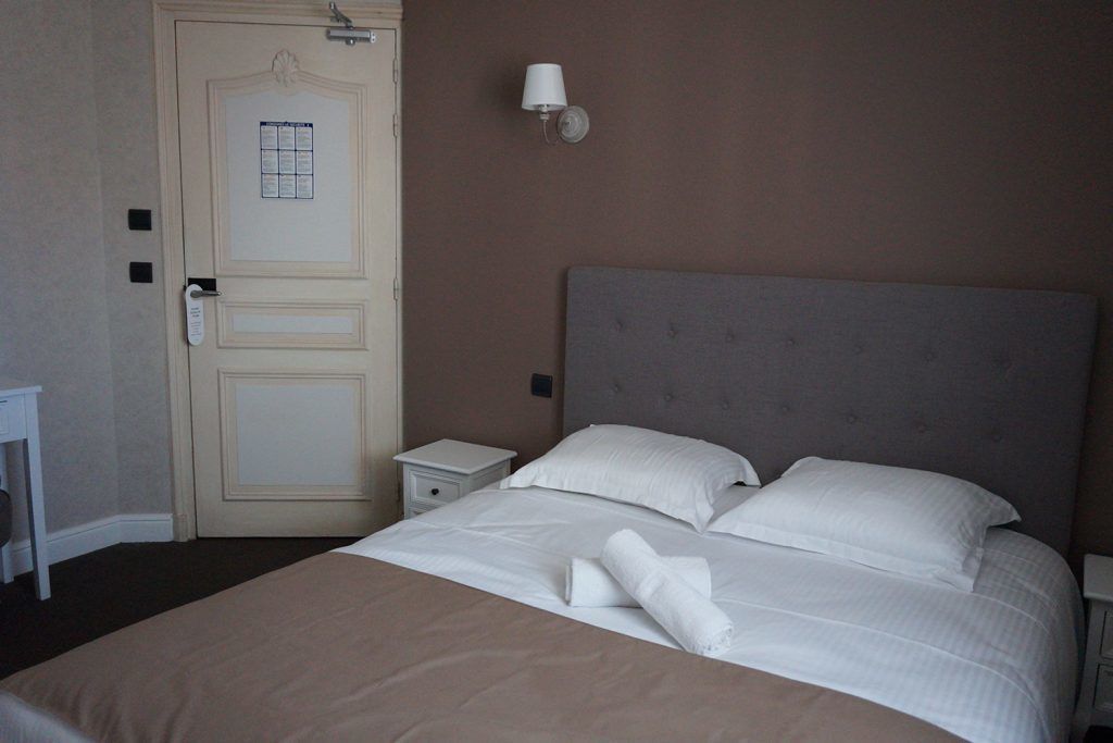 A hotel room with a large bed, a nightstand, a wall-mounted lamp, and a door with a bulletin board.