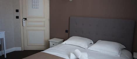 A hotel room with a large bed, a nightstand, a wall-mounted lamp, and a door with a bulletin board.