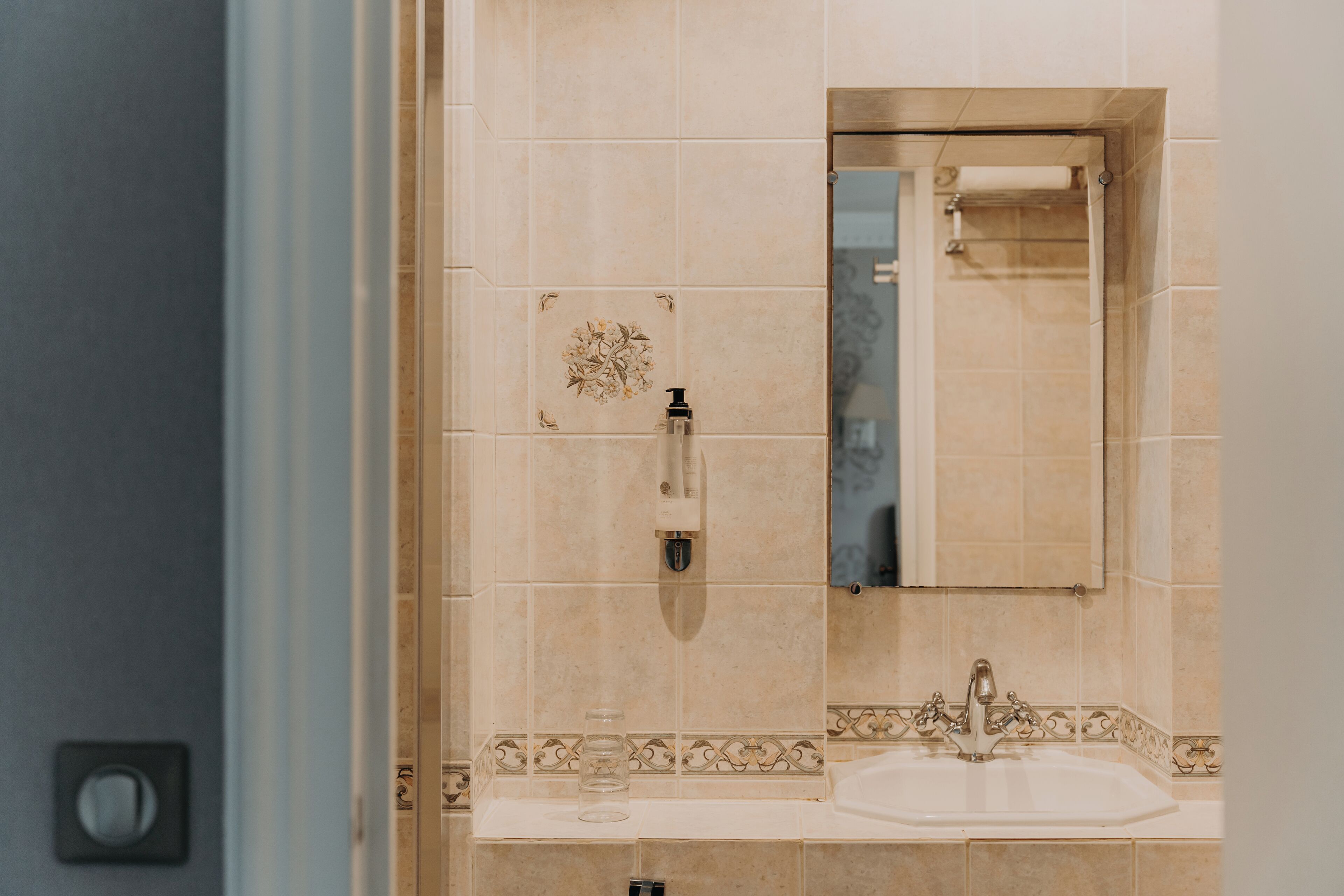 A bathroom with beige tiles, a sink, a mirror, and a soap dispenser.
