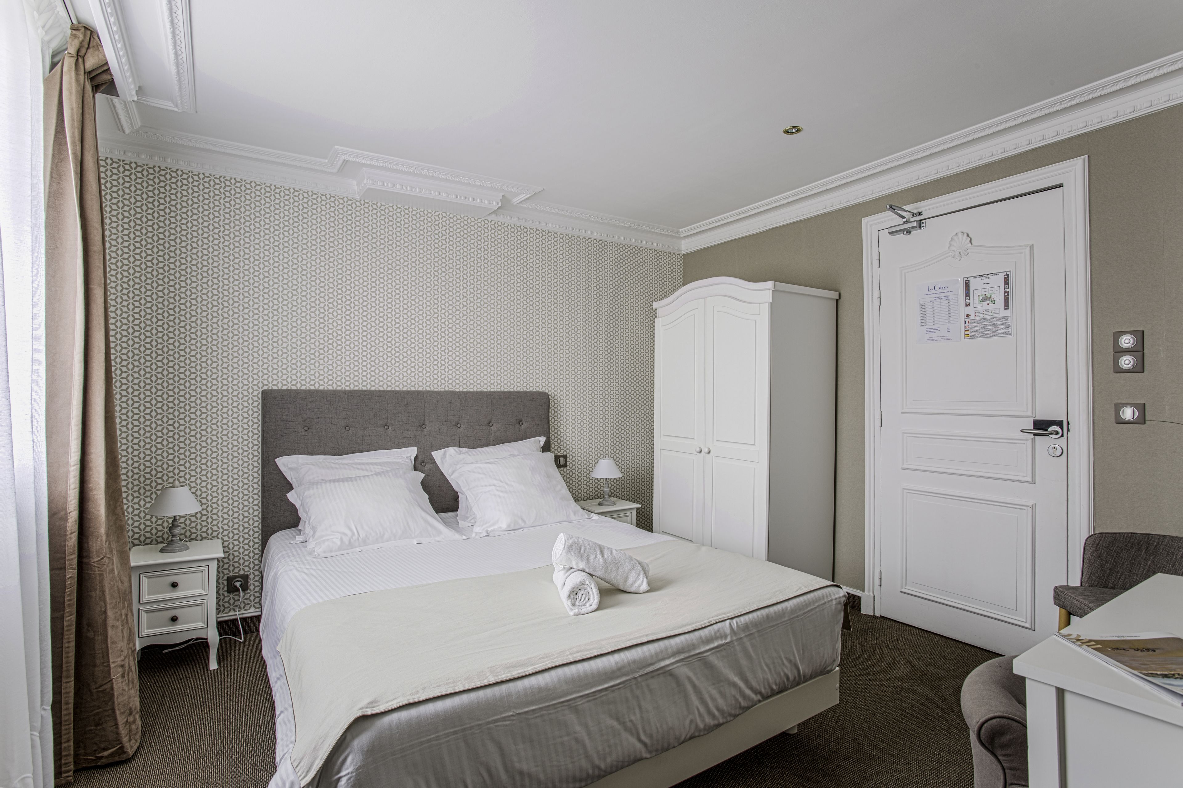 A neatly arranged hotel room with a large bed, a bedside table, a chair, a wardrobe, and a door with a lock and a notice.