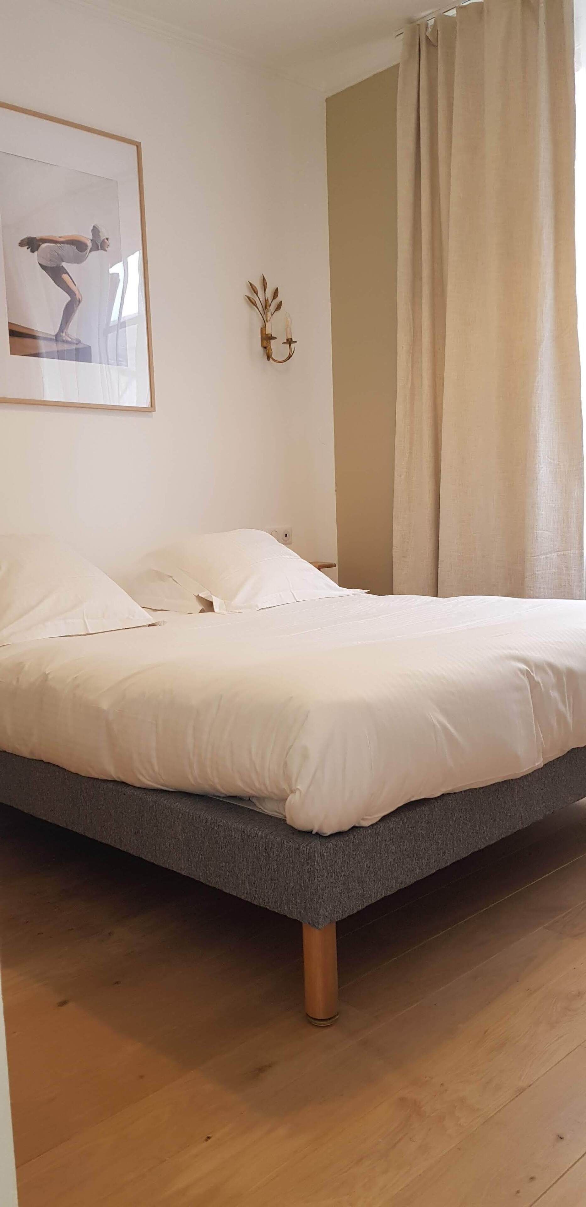 Una cama con ropa blanca, un cuadro en la pared y una cortina.