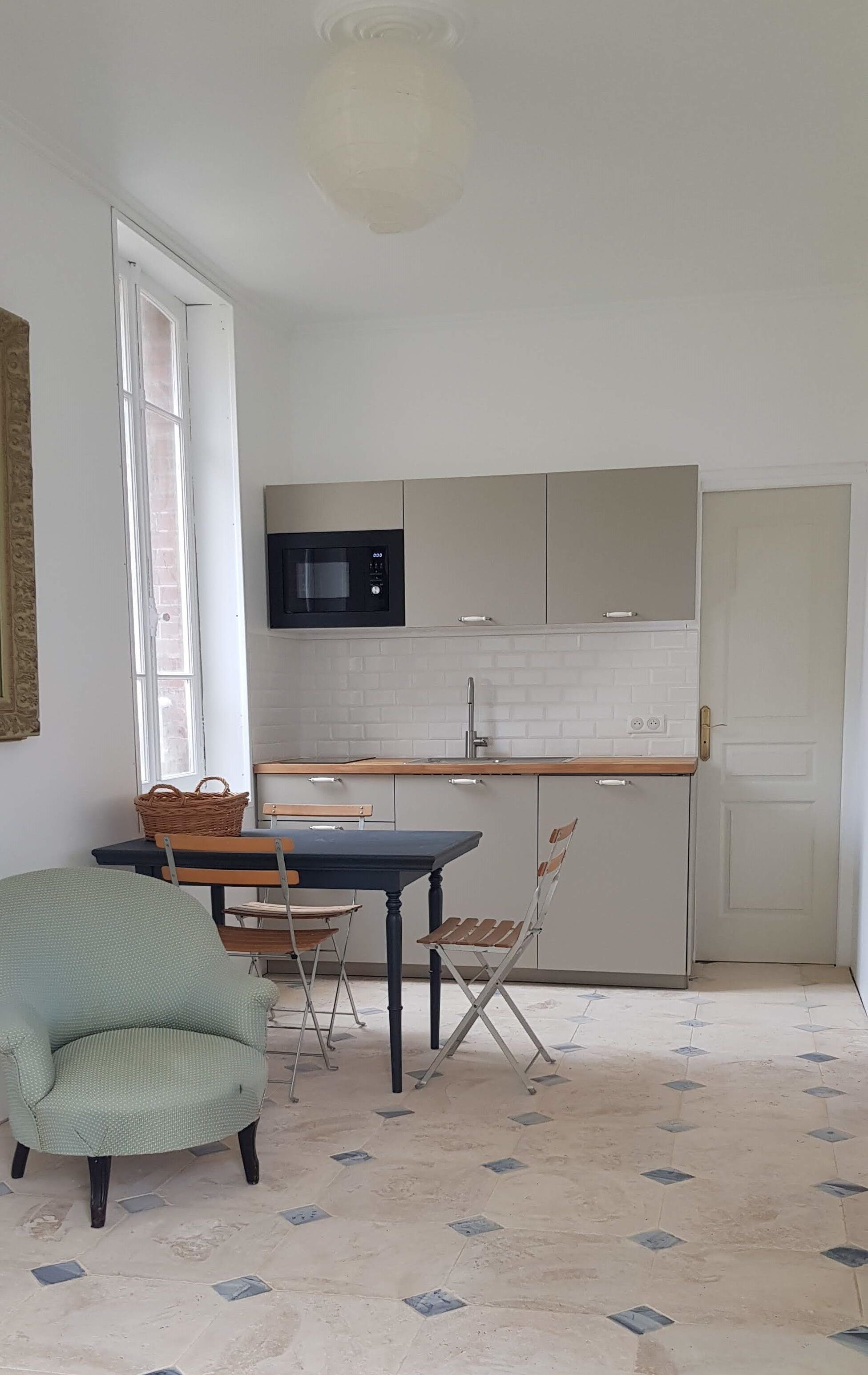Een kleine keuken met een magnetron, spoelbak en kastjes. Er staat een eettafel met stoelen en een rieten mand. De kamer heeft een betegelde vloer en een raam met uitzicht.