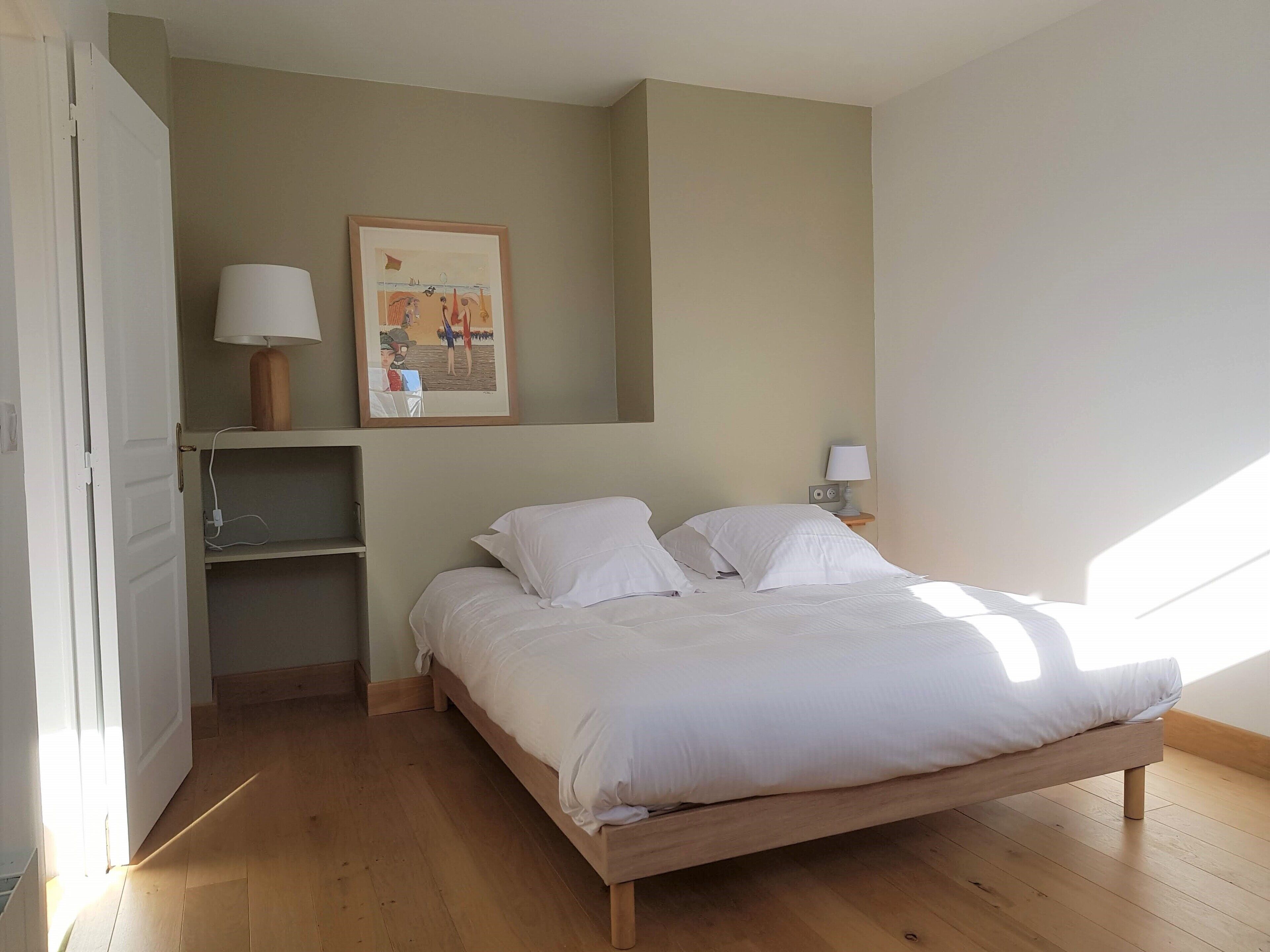 A bedroom with a wooden bed, a nightstand with a lamp, a shelf with a framed picture, and a door leading to another room.