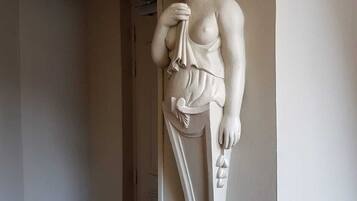 Una estatua de mármol blanco de una mujer sosteniendo un paño, ubicada sobre un pedestal en un pasillo con una puerta de madera y una pared blanca.